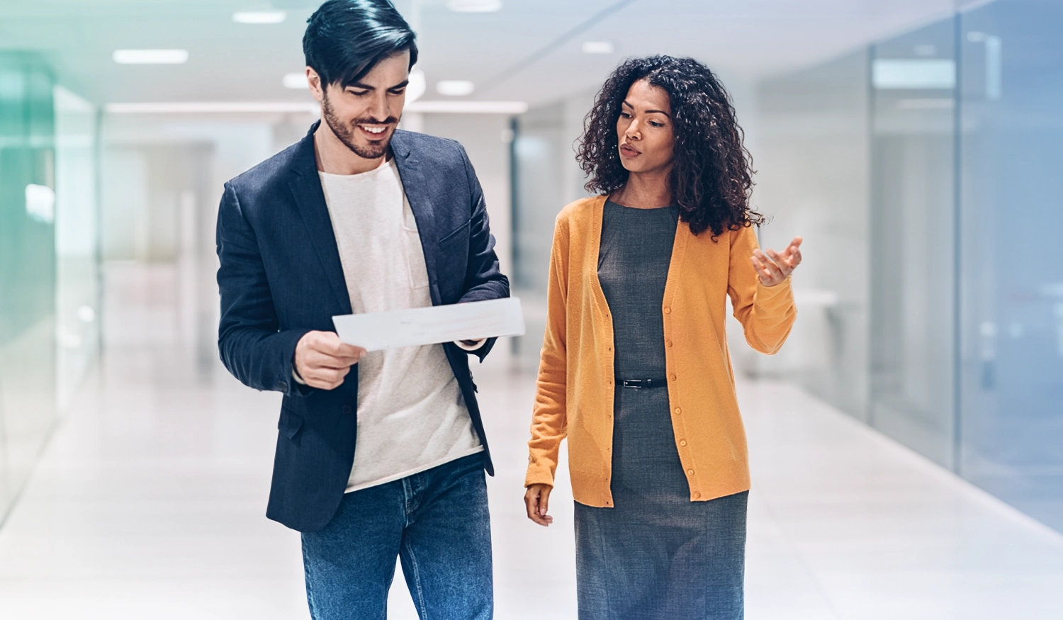 man in a tshirt and suit jacket and woman in a yellow cardigan walking down a white hallway looking a sheet of paper