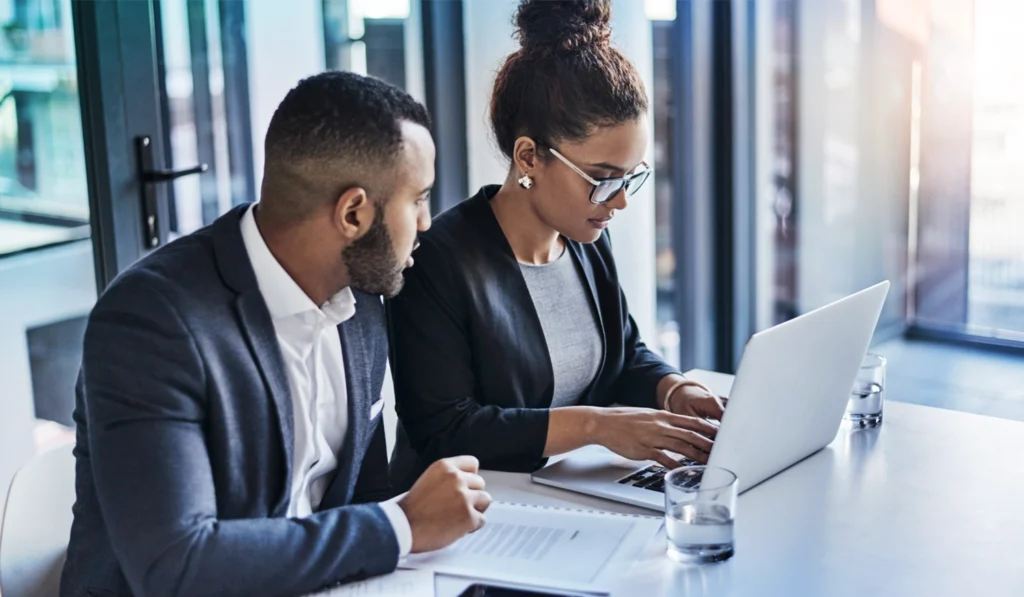 man and woman in business attire sitting at a white desk by a window looking at a computer together