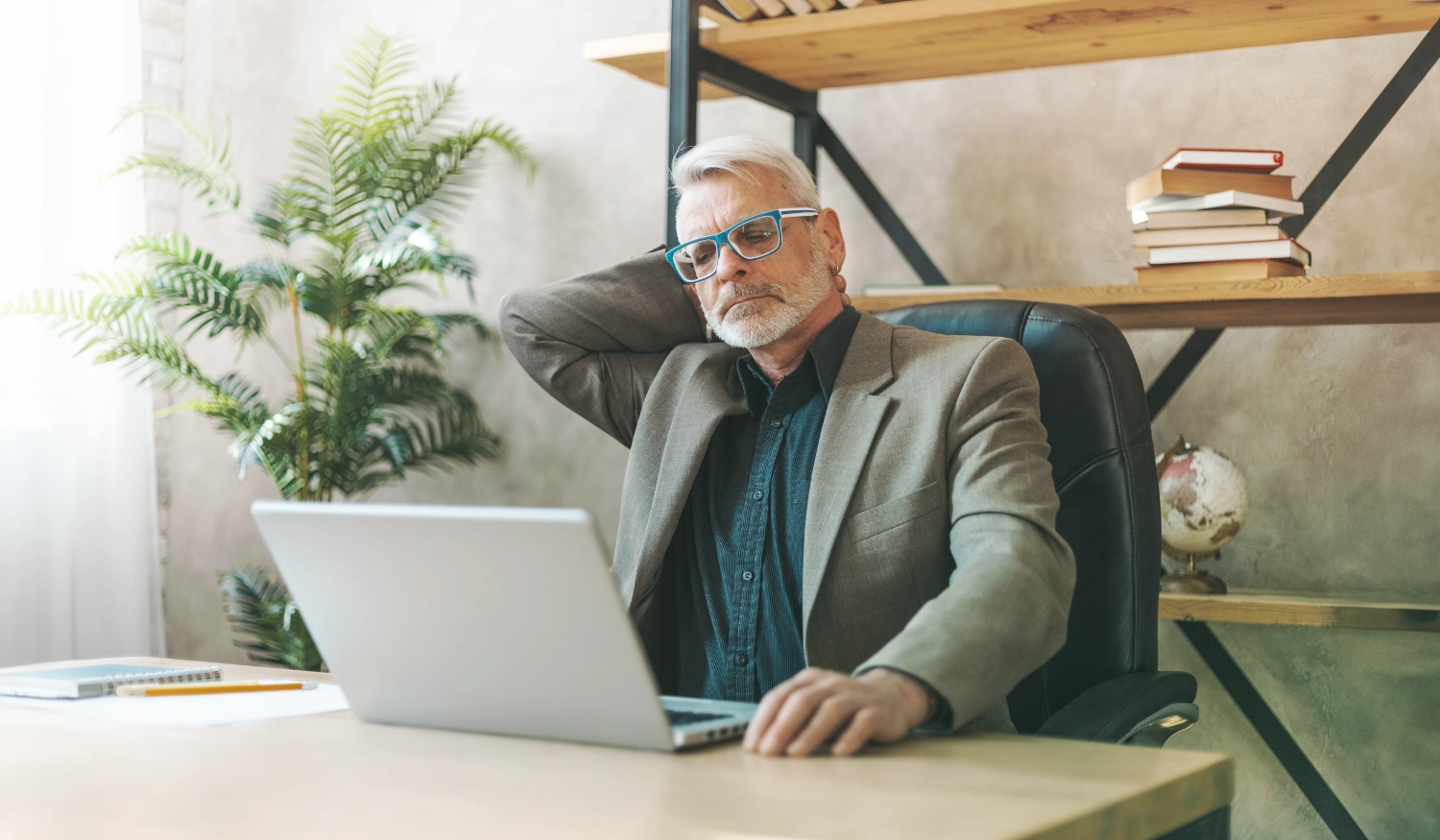man sitting in a chair at a desk looking at a laptop with bookshelf and plant behind