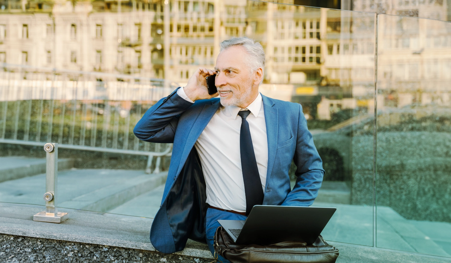 man sitting outside in a city wearing business attire on the phone with a laptop in his lap