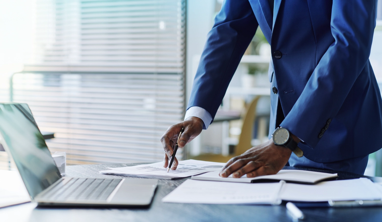 a man in an office standing at a desk with laptop and papers on it in a blue suit jacket